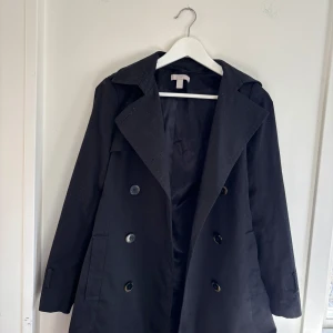 Svart kappa med knappar - Elegant svart kappa med dubbelknäppning och klassisk krage. Perfekt för våren med sin stilrena design och långa ärmar. Passar bra till både vardag och mer formella tillfällen.🩷