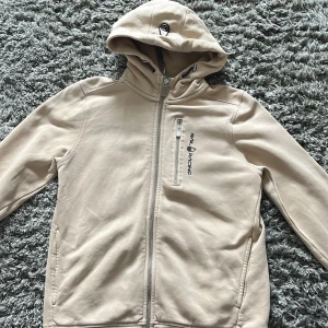 Beige jacka från Sail Racing - Snygg beige jacka från Sail Racing med huva och dragkedja. Jackan har en praktisk bröstficka med dragkedja och är perfekt för kyligare dagar. Den har en stilren design med logotyp på bröstet.