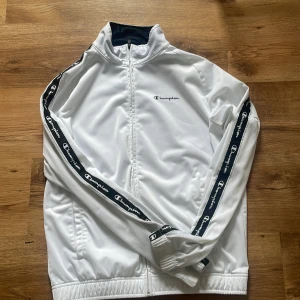 Vit träningsjacka från Champion - Säljer en snygg vit träningsjacka från Champion med dragkedja och logotyp längs ärmarna. Jackan har en hög krage och elastiska muddar för en bekväm passform. Perfekt för sportiga aktiviteter eller en avslappnad stil.