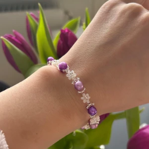 Lila och vitt pärlarmband - Charmigt armband med vita och lila pärlor i varierande nyans, dekorerat med små blommor. Perfekt för att ge en färgklick till din outfit. Armbandet har en justerbar kedja för perfekt passform. Gjord i äkta rocaillespärlor och silverpläterad kedja. 