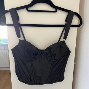 Svart korsettopp - Svart korsettopp. Toppen har en figurnära passform och är tillverkad i ett glansigt material. Det känns lite som satin vilket gör att den känns lyxig. Är knäppen bak att spänna med. 