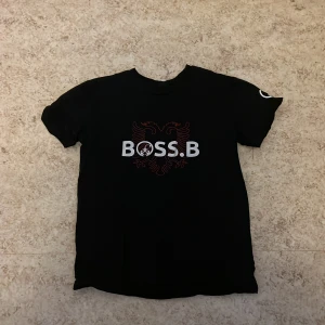 Svart t-shirt från Boss - Snygg svart t-shirt från Boss med trycket 'BOSS.B' och en stiliserad örn i rött. Perfekt för en casual look. Kortärmad och tillverkad i mjukt material för extra komfort.