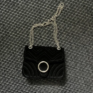 Svart axelväska i sammet - Snygg svart axelväska i sammet med kedjerem och ett stort, runt metallspänne på framsidan. Väskan har ett quiltat mönster som ger en lyxig känsla. Perfekt för att lyfta din outfit till nästa nivå!