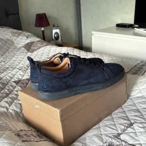Mörkblå sneakers från Christian Louboutin - Snygga mörkblå sneakers från Christian Louboutin med den ikoniska röda sulan. Skorna har en stilren design med snörning och är tillverkade i mocka. Perfekta för dig som vill ha en touch av lyx i vardagen.