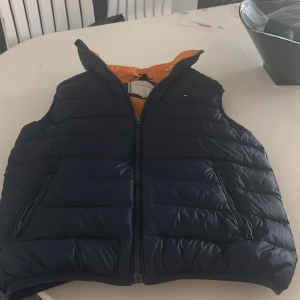 Svart dunväst från Tommy Hilfiger - Snygg svart dunväst från Tommy Hilfiger med orange foder. Västen har dragkedja framtill och två fickor med dragkedja. Perfekt för kyligare dagar och ger en stilren look. Storlek 152 i barn,pris kan sänkas vid snabb affär