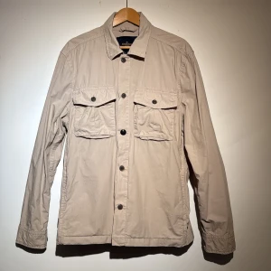 Beige Morris overshirt med knappar - Säljer en stilren beige overshirt med knappar framtill och två bröstfickor. Perfekt för lager-på-lager-stil och passar till många olika outfits. Enkelt och snyggt val för både vardag och mer uppklädda tillfällen.