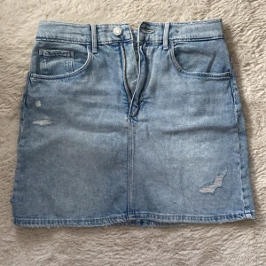 Ljusblå jeanskjol från H&M  - Säljer en fin ljusblå jeanskjol från H&M. Kjolen har en klassisk femficksdesign och slitna detaljer för en trendig look. Perfekt för en avslappnad stil. Den har en dragkedja och knapp framtill.