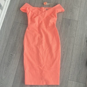 Korallfärgad klänning från River Island - Snygg korallfärgad klänning från River Island med offshoulder-design och dragkedja i ryggen. Klänningen har en figurnära passform och är tillverkad i ett ribbat material som ger en fin struktur. Perfekt för en somrig look!