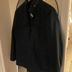 Mörkblå jacka från J.Lindeberg - Säljer en stilren mörkblå jacka från J.Lindeberg. Jackan har en diskret logotyp på armen och dragkedja vid kragen. Perfekt för en elegant look. Använd men i fint skick