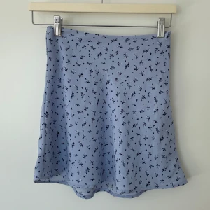 Blå blommig kjol från H&M - Söt ljusblå kjol med ett fint blommigt mönster i mörkare nyanser. Kjolen har en kort längd och är perfekt för varmare dagar. Lappen är bortklippt men är i storlek 32/XXS och är från H&M divided. Dragkedja i sidan och är använd fåtal gånger. 💕