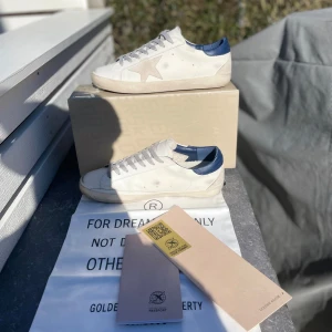Vita sneakers med blå detaljer från Golden Goose - Snygga vita sneakers från Golden Goose med en ikonisk stjärndetalj på sidan. Skorna har blå detaljer vid hälen och grå snörning. Perfekta för en stilren och avslappnad look.