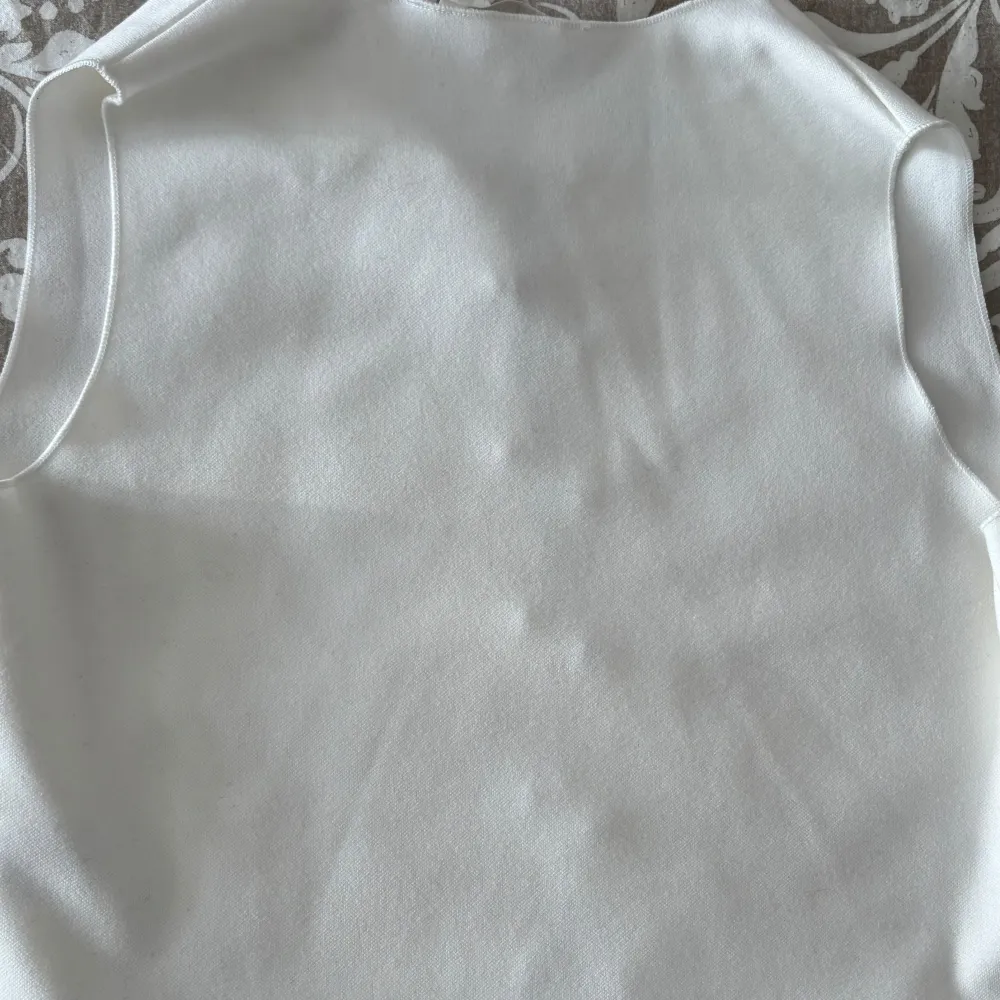 Elegant vit väst med fem guldknappar framtill. Perfekt för att ge en stilren touch till din outfit. Västen har en klassisk design och är tillverkad i ett mjukt material.. Puvut & Bleiserit.
