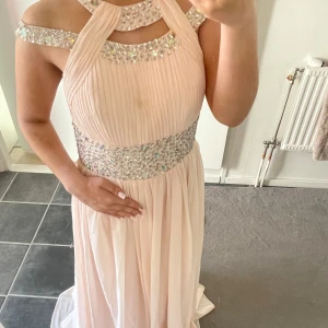 Ljusrosa långklänning med strassdetaljer - Elegant ljusrosa långklänning med glittrande strassdetaljer på axelband och midja. Klänningen har en unik halterneck-design och en rygg med snörning. Perfekt för speciella tillfällen. Har små omärkbara defekter därav priset, men det är ingenting man märker/tänker på. Vet ej den exakta storleken men den passar mig som bär allt mellan storlekarna Xs, S och M så någonstans där imellan. 