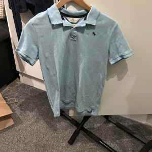 Ljusblå pikétröja från H&M - Säljer en ljusblå pikétröja från H&M med klassisk krage och korta ärmar. Tröjan har en liten broderad logga på bröstet och är perfekt för en avslappnad stil. Storleken är 164