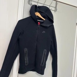 Svart hoodiejacka från Nike - Svart hoodiejacka från Nike med dragkedja framtill och två fickor med dragkedja. Jackan har huva och diskret Nike-logga på bröstet. Perfekt för en sportig och avslappnad stil. Gillar inte den är inte så mke använd helt ny är xs passar S 