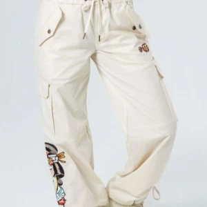 Beige cargobyxor med broderier från Ed Hardy - Nyskick! SX! Snygga beige cargobyxor från Ed Hardy med broderad geisha på benet och stora fickor. Byxorna har dragsko i midjan och Ed Hardy-logga i rött på baksidan. Perfekta för dig som vill sticka ut med coola detaljer.
