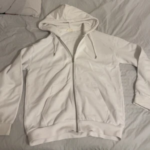 Vit hoodie med dragkedja - Säljer en stilren vit hoodie med dragkedja och huva. Tröjan har långa ärmar, två fickor framtill och ribbade muddar vid ärmslut och nederkant. Perfekt för en avslappnad look. Tröjan är från Pier One (aldrig använt)