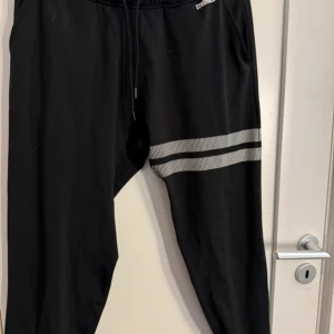 Svarta mjukisbyxor med vita ränder - Svarta mjukisbyxor med två vita ränder över ena låret. Byxorna har dragsko i midjan och är perfekta för en avslappnad stil. Materialet känns mjukt och bekvämt, och passformen är normal.