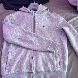 Grå hoodie från Lacoste Sport - Säljer en grå hoodie från Lacoste Sport med dragkedja och huva. Tröjan har två fickor framtill och den klassiska Lacoste-loggan på bröstet. Perfekt för en sportig och avslappnad stil.