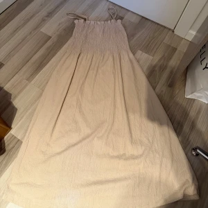 Beige långklänning med smock och tunna axelband - Säljer en beige långklänning med smock upptill och tunna axelband. Klänningen har en luftig och flowy passform, perfekt för varma dagar. Materialet känns mjukt och har en lätt struktur. Enkel och stilren design som passar till många tillfällen.
