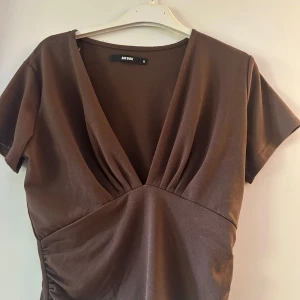 Brun omlottopp från Bik Bok - Säljer en brun omlottopp från Bik Bok med djup v-ringning och korta ärmar. Toppen har rynkade detaljer i sidorna och är tillverkad i ett mjukt, stretchigt material. Perfekt för dig som gillar en stilren och trendig look. 