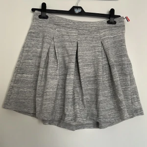 Grå kjol från Tommy Hilfiger - Säljer en grå kortkjol från Tommy Hilfiger med veck framtill och dragkedja i sidan. Kjolen har en mjuk och stretchig känsla, perfekt för en avslappnad men ändå stilren look.