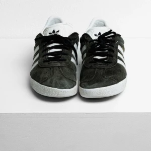Adidas Gazelle sneakers i svart mocka - Säljer ett par klassiska Adidas Gazelle sneakers i svart mocka med vita detaljer och vita ränder på sidorna. Skorna har snörning och vit sula, samt Adidas-logga på häl och plös. Perfekta för en stilren vardagslook. Skorna är använda väldigt lite, som nya. Nypris 1000 kr, pris kan diskuteras
