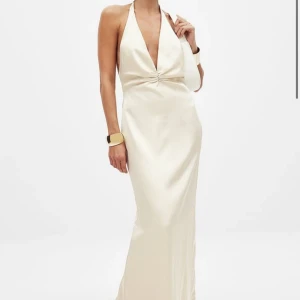 Champagnefärgad maxiklänning med halterneck - Elegant långklänning i champagnefärgat satinmaterial med halterneck. Klänningen har en slät och glansig yta som ger en lyxig känsla och faller snyggt längs kroppen. Perfekt för speciella tillfällen som t.ex. balen. ALDRIG använd! Helt slutsåld 