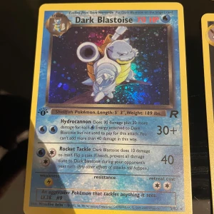 Pokémonkort Dark Blastoise Holo - Säljer ett original Pokémonkort: Dark Blastoise Holo från Team Rocket-setet. Kortet har en blå ram, holografisk bild och engelska texter. Motivet visar Blastoise med kanoner på ryggen. Perfekt för samlare av Pokémonkort!