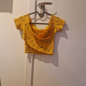 Gul croppad topp med blommönster - Säljer en söt gul croppad topp med små blommor. Toppen har korta ärmar och en volangdetalj framtill. Perfekt för varma dagar och ger en somrig känsla.