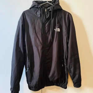 Svart vindjacka från The North Face - Säljer en svart vindjacka från The North Face med huva och dragkedja framtill. Jackan har två fickor med dragkedja och justerbar huva. Perfekt för blåsiga dagar och enkel att packa ner i väskan.