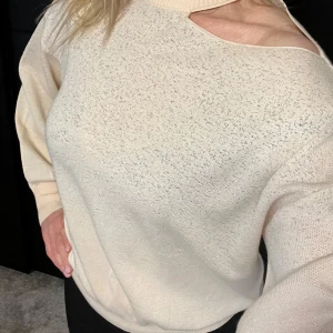 Beige stickad tröja med oneshoulder-detalj - Säljer en beige stickad tröja med unik oneshoulder-detalj och ribbad hög krage. Tröjan har lång ärm och avslappnad passform, perfekt för dig som vill ha något trendigt och bekvämt.