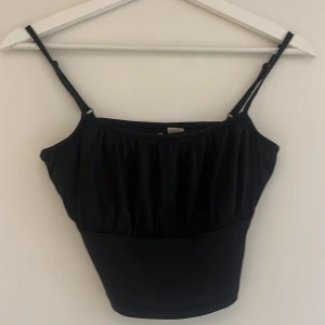 Svart croppad linnetopp från Cubus - Säljer en svart croppad linnetopp från Cubus med smala axelband och lätt rynkad detalj framtill. Toppen har en enkel och stilren design, perfekt att matcha med jeans eller kjol. Justerbara axelband för bästa passform.