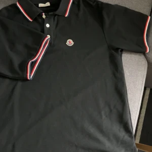 Svart pikétröja från Moncler - Säljer en svart pikétröja från Moncler med klassisk krage och knappar framtill. Tröjan har Moncler-logga på bröstet och röda, vita och blå ränder på krage och ärmslut. Perfekt för en stilren och sportig look.