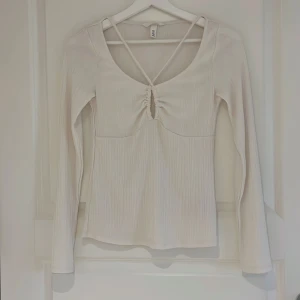 Blus - Säljer en vit långärmad topp från H&M med ribbad struktur och snygg knytdetalj framtill. Toppen har djup urringning och smala band som korsas i ryggen. Perfekt för en trendig och stilren look.