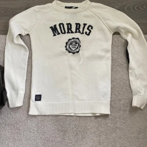 Vit stickad tröja från Morris - Vit stickad tröja från Morris med svart broderad logga och emblem på bröstet. Tröjan har rund halsringning och långa ärmar med ribbade muddar och nederkant. Perfekt för en stilren och klassisk look. Skick: 7/10 Storlek: S