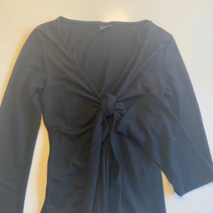 Svart långärmad topp med knyt från Gina Tricot - Säljer en svart långärmad topp från Gina Tricot med snygg knytning framtill. Toppen har en djup v-ringning och är tillverkad i ett mjukt material som sitter snyggt på kroppen. Perfekt att styla med jeans eller kjol.