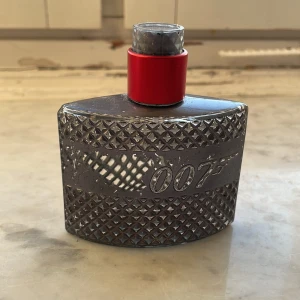 James Bond 007 Eau de Toilette - Maskulin parfym med aromatiska och fräscht kryddiga toner, inslag av trä, läder och citrus. Flaskan är silverfärgad med diamantmönster och röd hals, prydd med 007-loggan.