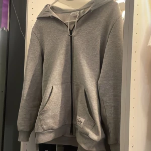 Grå hoodie med dragkedja från Andy by Frank Dandy - Säljer en enkel och stilren grå hoodie från Andy by Frank Dandy. Tröjan har dragkedja framtill, två fickor och huva. 