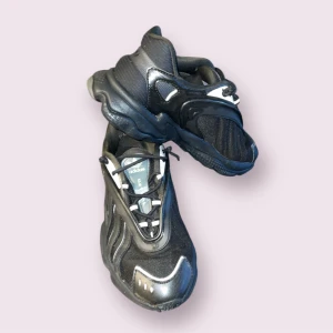 Svarta sneakers från Adidas - Säljer ett par svarta Adidas sneakers med futuristisk design och reflekterande detaljer. Skorna har snörning, rund tå och platt sula. Perfekta för dig som gillar en sportig och modern stil.