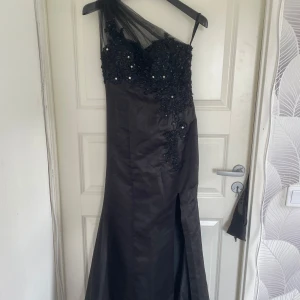 Svart långklänning med spets och paljetter - Säljer en svart långklänning med en axel och transparent tyll. Överdelen är dekorerad med spets, paljetter och pärlor. Klänningen har slits framtill och en elegant, figurnära passform. Perfekt för bal eller fest.
