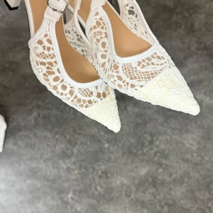 Vita spetsiga pumps med spets och klack - Säljer ett par vita pumps med spetsig tå och klack. Skorna har fina spetsdetaljer och en justerbar rem runt ankeln. Perfekta för dig som vill ha något elegant och unikt till din outfit. Helt nya är tyvärr för stora för mig därav säljs dom, har en super bekväm passform med inbyggt stöd 