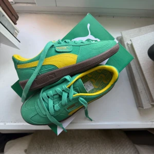 Grön/gula sneakers från Puma - Säljer ett par snygga Puma sneakers i grönt med gula detaljer och brun sula. Skorna har klassisk snörning och är tillverkade i mocka med textilfoder. Perfekta för dig som vill sticka ut med färg på fötterna!