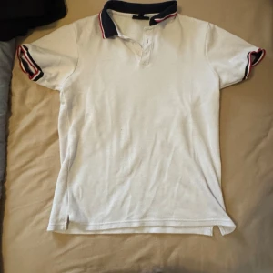 Vit pikétröja med shorts från SMOG - Säljer en klassisk vit pikétröja med shorts från  SMOG i storlek S. Tröjan har korta ärmar, krage i mörkblått med röda och vita ränder samt knappar vid halsen. Perfekt för en stilren och sportig look.