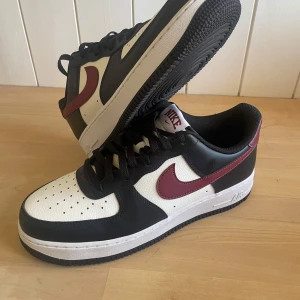 Nike Air Force 1 med vinröd swoosh EUR 44 - Helt oanvända Nike Air Force 1 sneakers med svart och vit bas samt vinröd swoosh och häl. Klassisk design med rund tå, snörning och platt sula. Storlek 44, bara att skriva vid minsta lilla fundering.