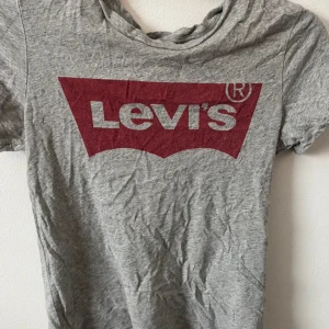 Grå t-shirt från Levi's med röd logga - Säljer en klassisk grå t-shirt från Levi's med den ikoniska röda loggan framtill. T-shirten har rund halsringning och korta ärmar. Perfekt till jeans eller shorts för en avslappnad look.Frakt tillkommer