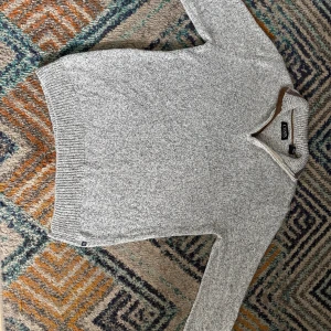 Grå stickad tröja med dragkedja från Chaps - Säljer en gråmelerad stickad tröja från Chaps med halv dragkedja och hög krage. Tröjan har ribbade muddar och en snygg detalj i mockaimitation vid dragkedjan. Perfekt för kyliga dagar.