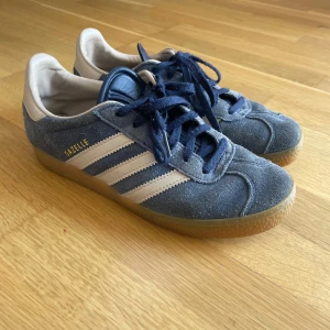 Adidas Gazelle blå sneakers - Säljer ett par klassiska Adidas Gazelle sneakers i blå mocka med vita detaljer och gummisula. Skorna har snörning och de ikoniska tre ränderna på sidan samt Gazelle-logga i guld. 