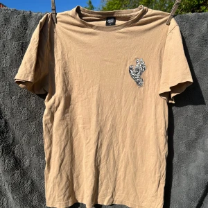 Beige t-shirt med svart tryck från Santa Cruz strl M - Beige t-shirt med stor svart och vit hand-illustration på ryggen och mindre tryck på bröstet. Rund halsringning och korta ärmar. Perfekt för dig som gillar streetwear och unika prints. Fint skick! Strl M