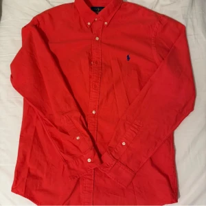 Röd skjorta från Ralph Lauren - Klassisk röd skjorta från Ralph Lauren med blå broderad logga på bröstet. Skjortan har lång ärm, knappar framtill och button-down krage. Tillverkad i bomull för en skön känsla och snygg passform.